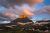 Golden sunlit summit rises above snow patches and green meadows under a pink cloud-filled sky with a moon at Glacier National Park.