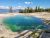 Turquoise geothermal pool with green edges near Yellowstone National Park, beside mineral crust and pine-covered lakefront shore.
