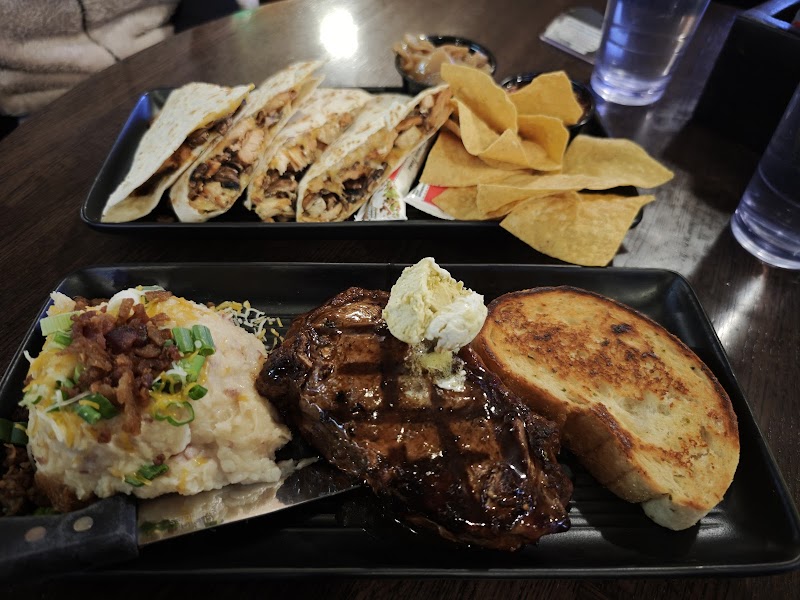 Grilled steak with butter, mashed potatoes topped with bacon and chives, and toasted bread on a black tray, with tacos and tortilla chips on a second tray in Yellowstone National Park.