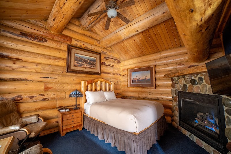 Cozy log cabin bedroom in Yellowstone National Park with a white bed, stone fireplace, wood walls, and landscape art.