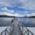 A wooden pier extends into Lake McDonald at Apgar Beach, Glacier National Park, with snow-capped peaks in the distance.