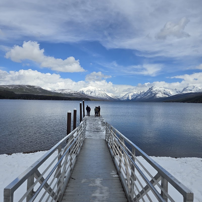 A wooden pier extends into Lake McDonald at Apgar Beach, Glacier National Park, with snow-capped peaks in the distance.