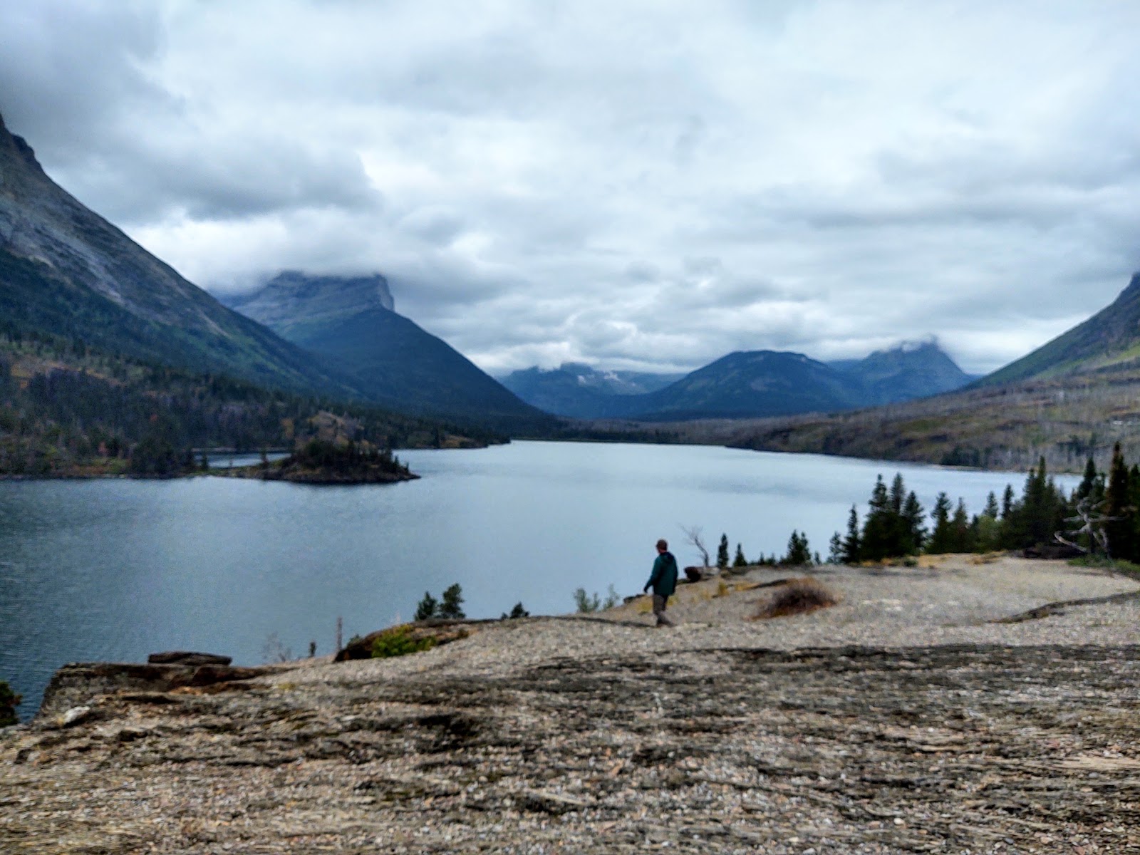 Rocky shore and calm glacier lake with distant mountains and a hiker near Glacier National Park