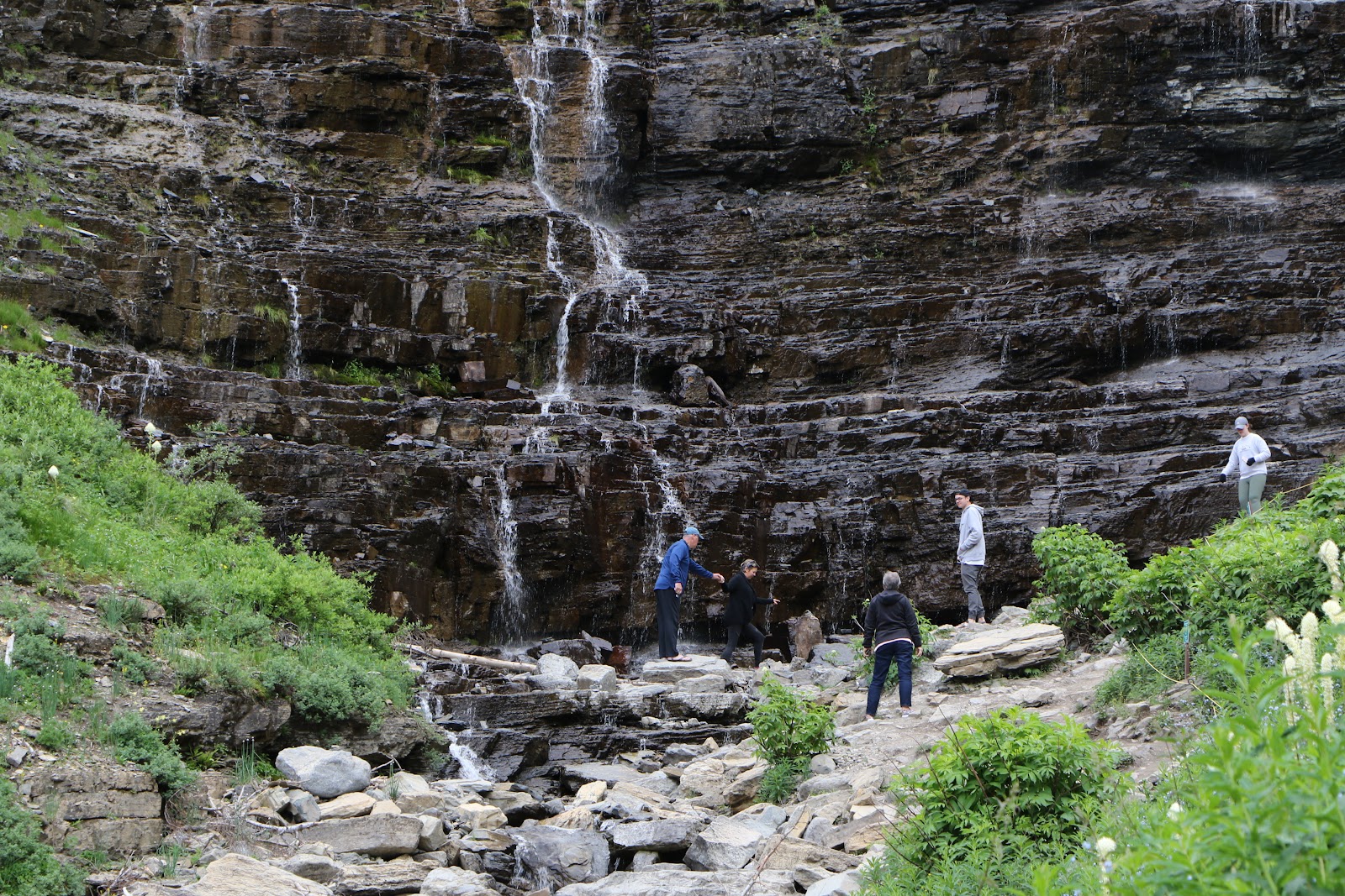 Going-to-the-Sun Mountain area in Glacier National Park features a rugged rock face with multiple small waterfalls and hikers along a rocky ledge.