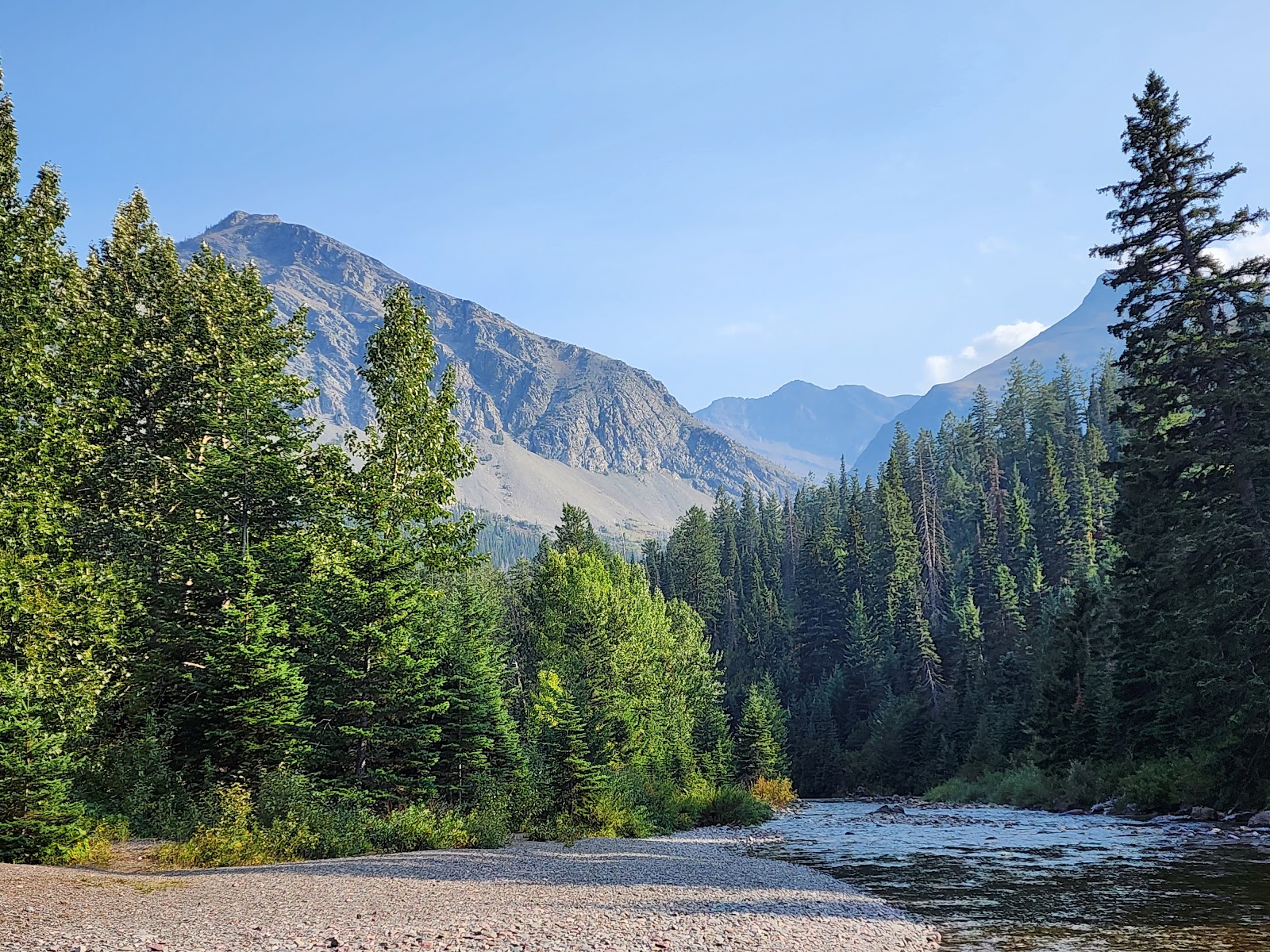 Running Eagle Falls Nature Trail in Glacier National Park winds along a river with green pines and rugged, rocky mountains in the distance.