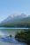 Rainbow Peak rises above a turquoise Glacier National Park lake, surrounded by evergreen forest and a clear blue sky.