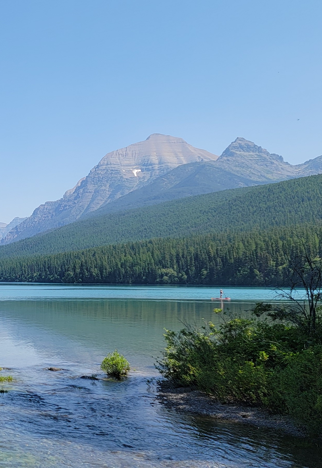 Rainbow Peak rises above a turquoise Glacier National Park lake, surrounded by evergreen forest and a clear blue sky.