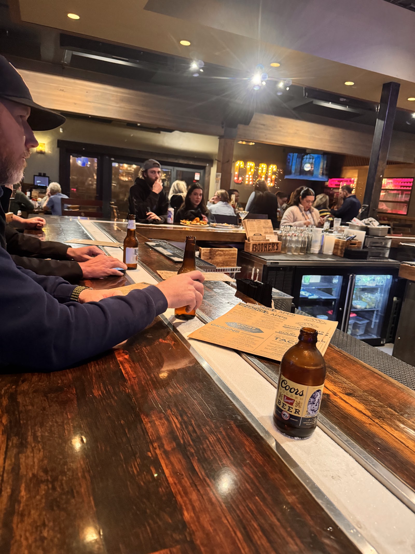 Inside a rustic Glacier National Park bar with a wooden counter and patrons enjoying drinks.