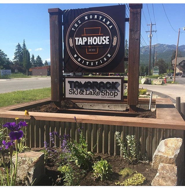 Roadside sign near Glacier National Park for a Montana tavern and ski shop with flowers in front.