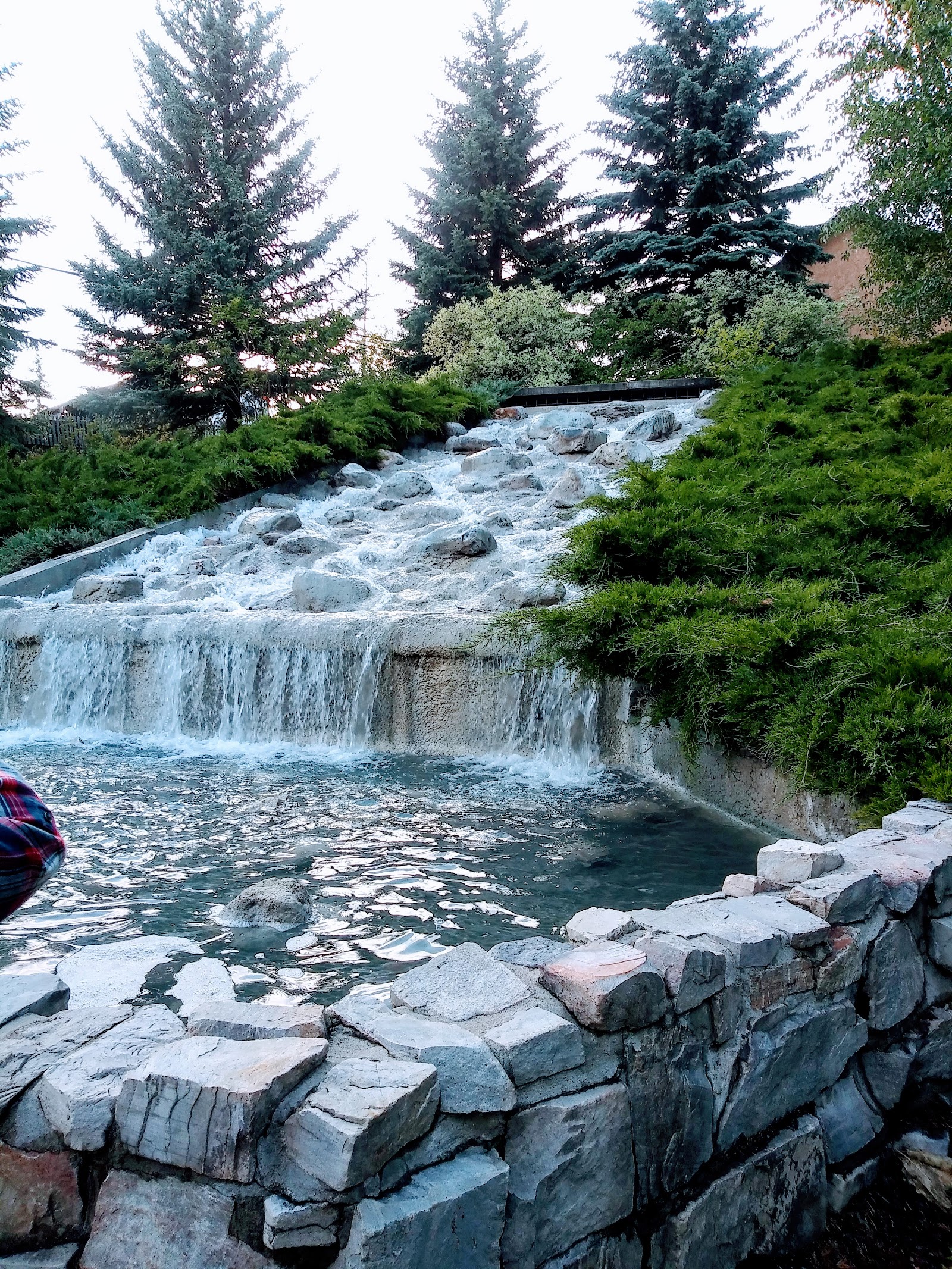 Stone-lined cascade over a low spillway into a rocky pool, with green shrubs and tall evergreens in Glacier National Park.