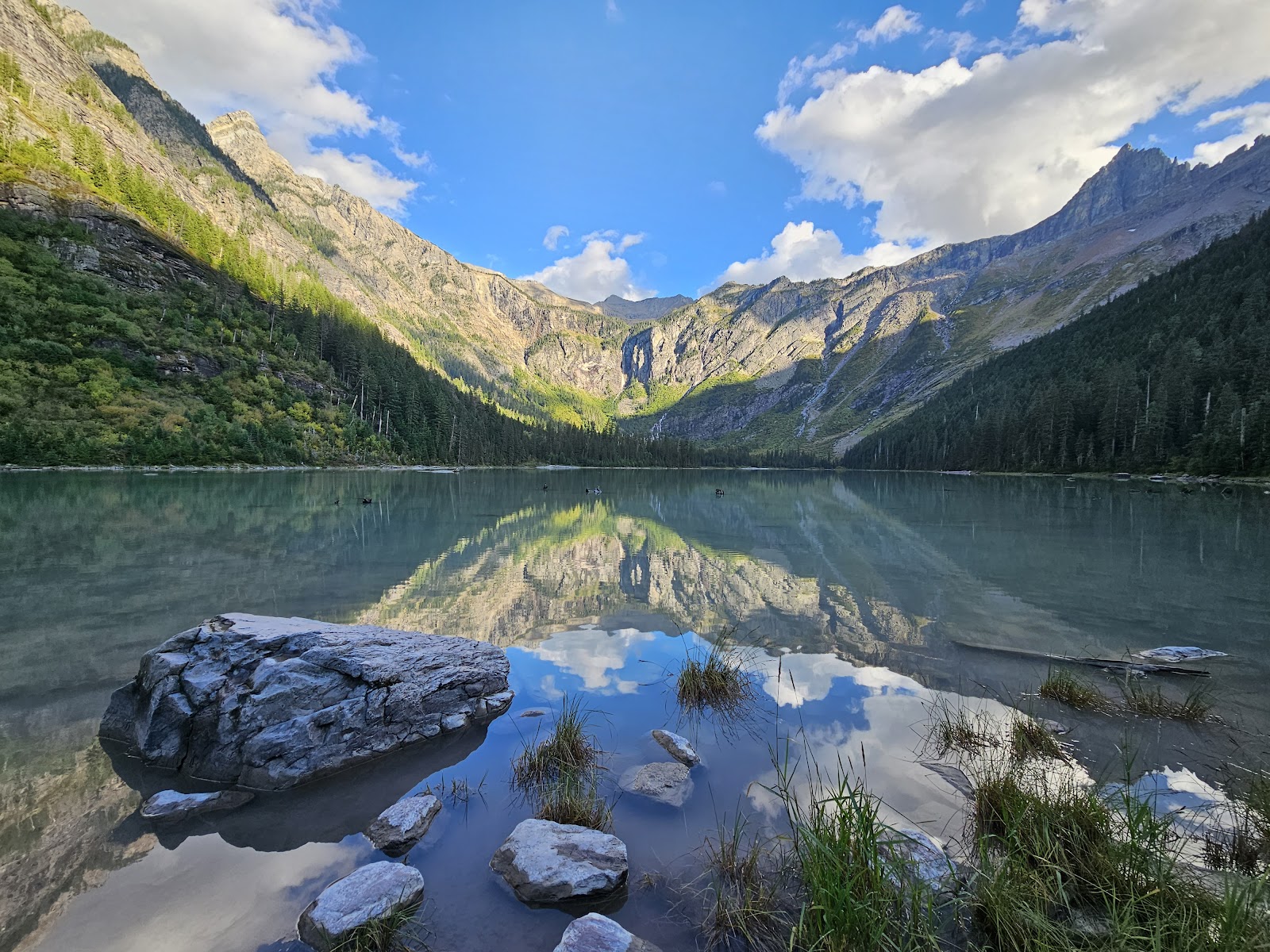 Trail of the Cedars Nature Trailhead at Glacier National Park, a tranquil lakeside scene with forested slopes and rugged peaks.