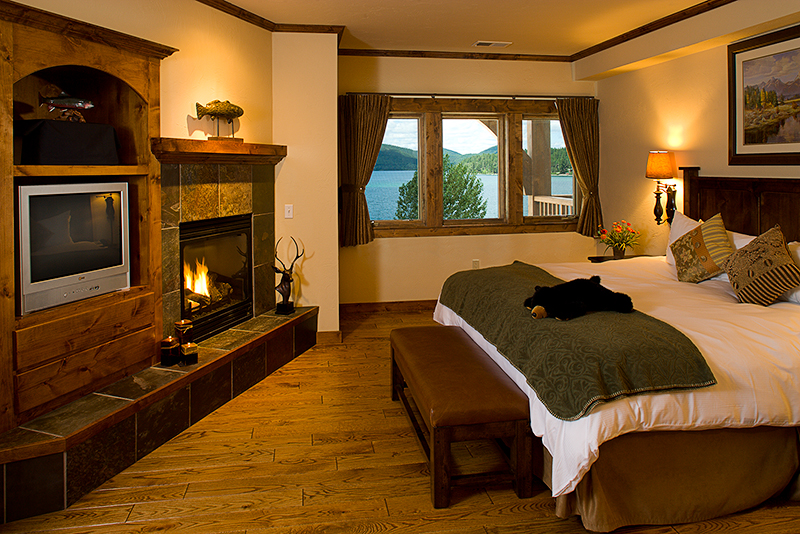 Cozy lodge bedroom with a stone fireplace and a large window overlooking a lake and mountains in Glacier National Park.