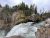 Rushing whitewater drops over a rocky brink, with pine trees and mossy cliffs at Yellowstone National Park.