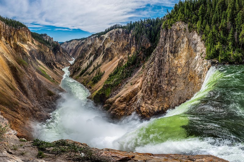 Yellowstone National Park — steep, colorful canyon walls frame a green river plunging over a waterfall with mist rising.