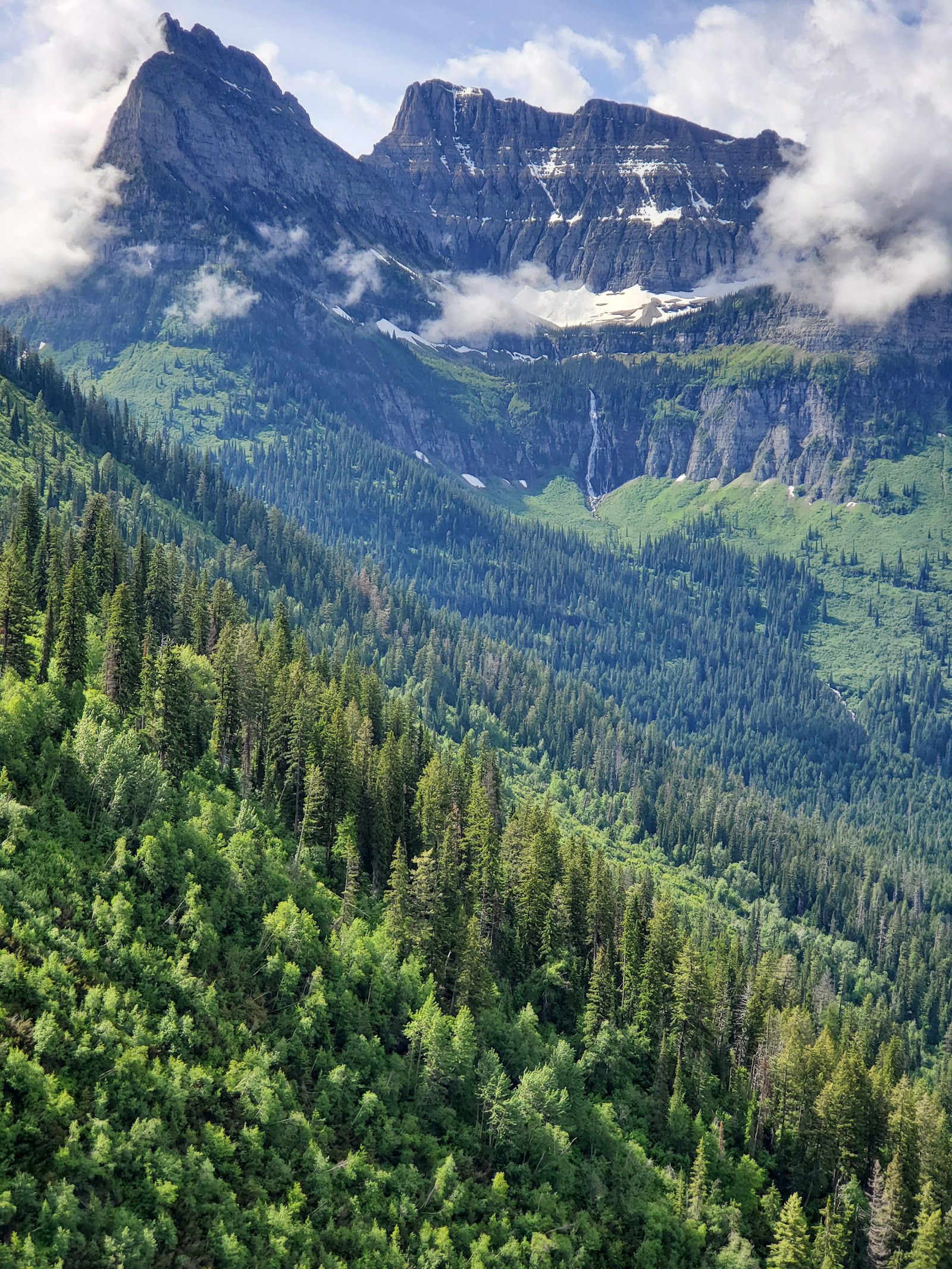 Rugged mountain peak above a lush evergreen valley in Glacier National Park, with a thin waterfall and clouds.