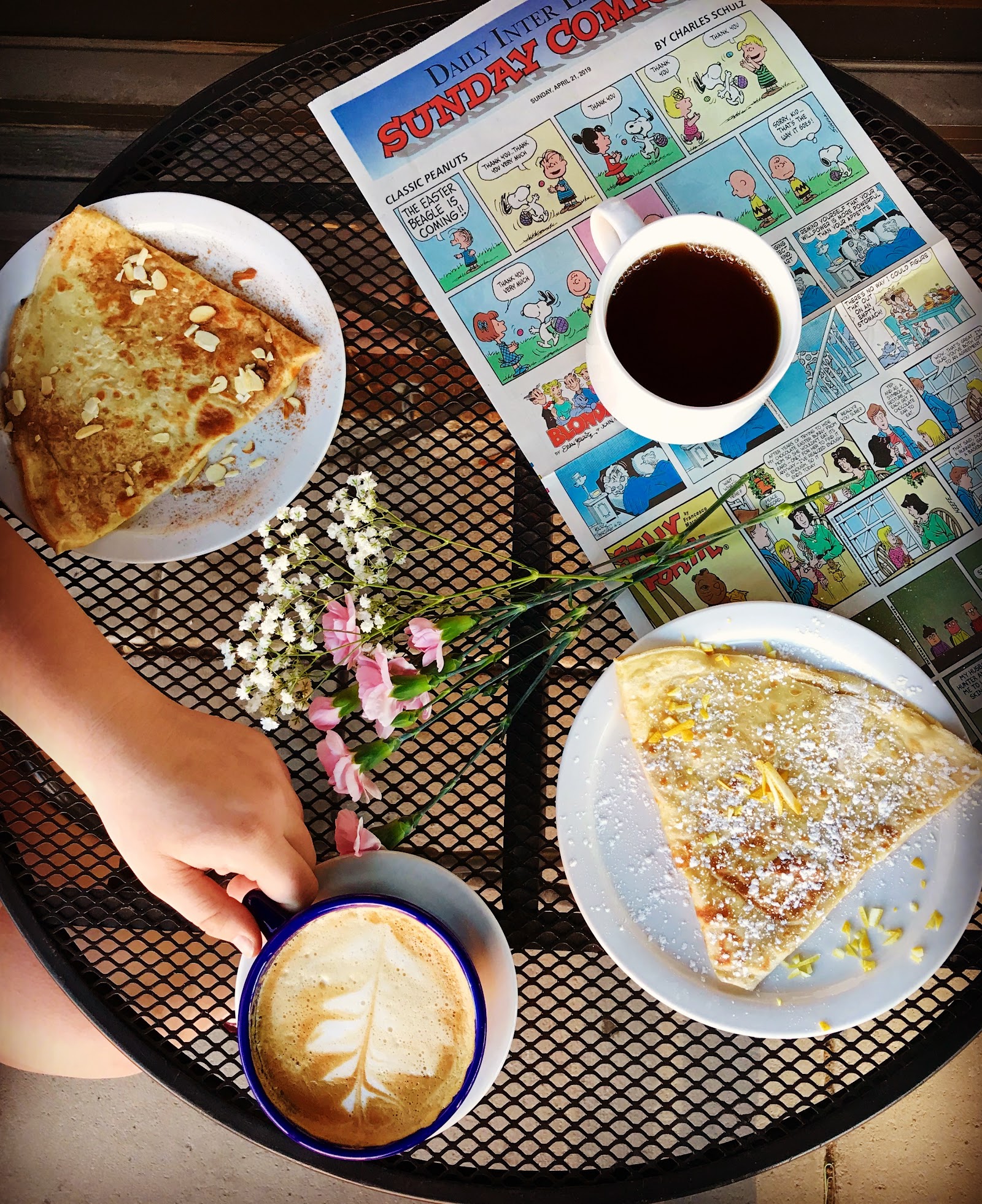 Crepe cafe breakfast with two crepes and coffee on a metal table at Glacier National Park, outdoors near a small floral arrangement.