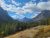 Redrock Falls stands in Glacier National Park with evergreen forests, rugged peaks, and a bright blue sky above the grassy foreground.