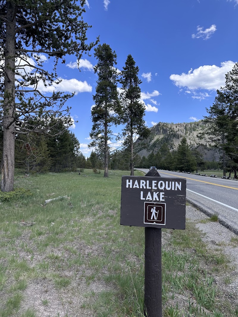 Yellowstone National Park trailhead sign for Harlequin Lake beside a road, with pines, grassy field, and distant mountains.