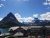 Jagged, snow-dusted peaks loom over Swiftcurrent Lake, with a dark lodge and parked cars along the shore in Glacier National Park.