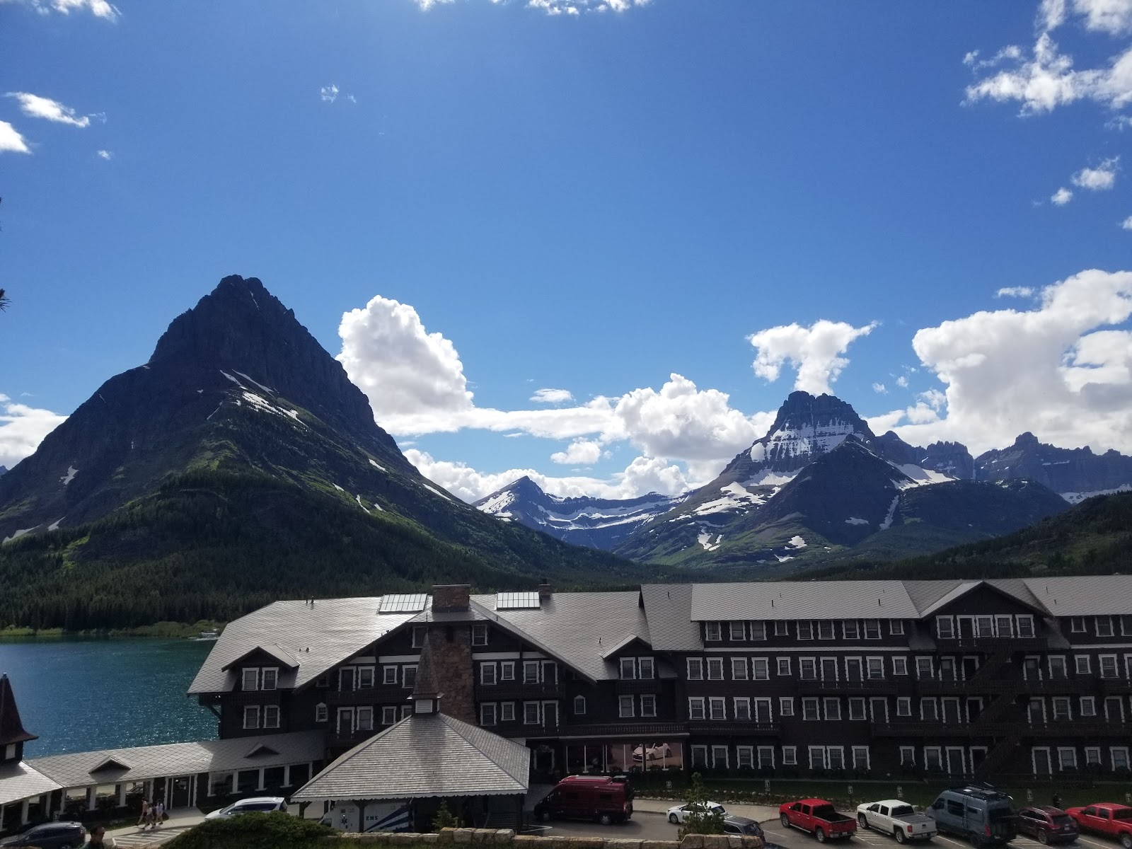 Jagged, snow-dusted peaks loom over Swiftcurrent Lake, with a dark lodge and parked cars along the shore in Glacier National Park.