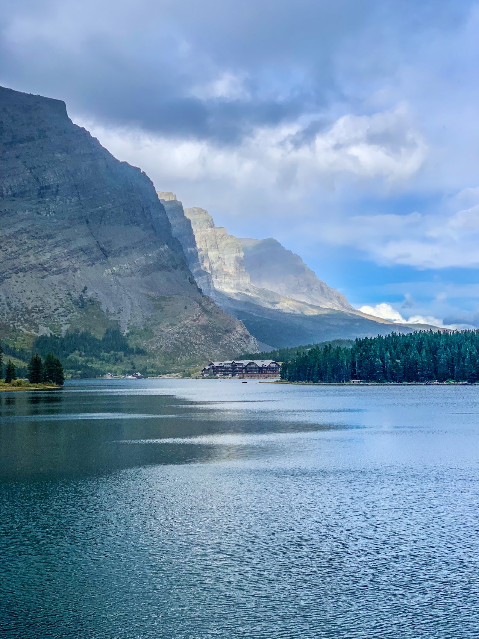 Swiftcurrent Lake reflects towering cliffs and evergreen forest beneath a blue, cloudy sky in Glacier National Park.