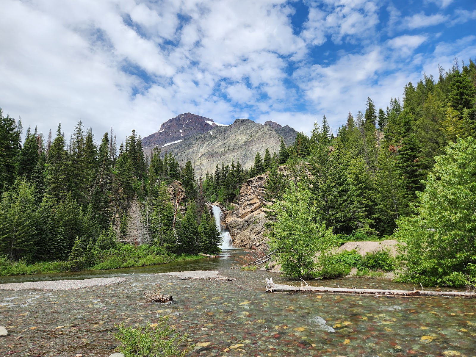 Running Eagle Falls cascades down a rocky ledge into a clear river, surrounded by evergreen pines and distant mountains in Glacier National Park.
