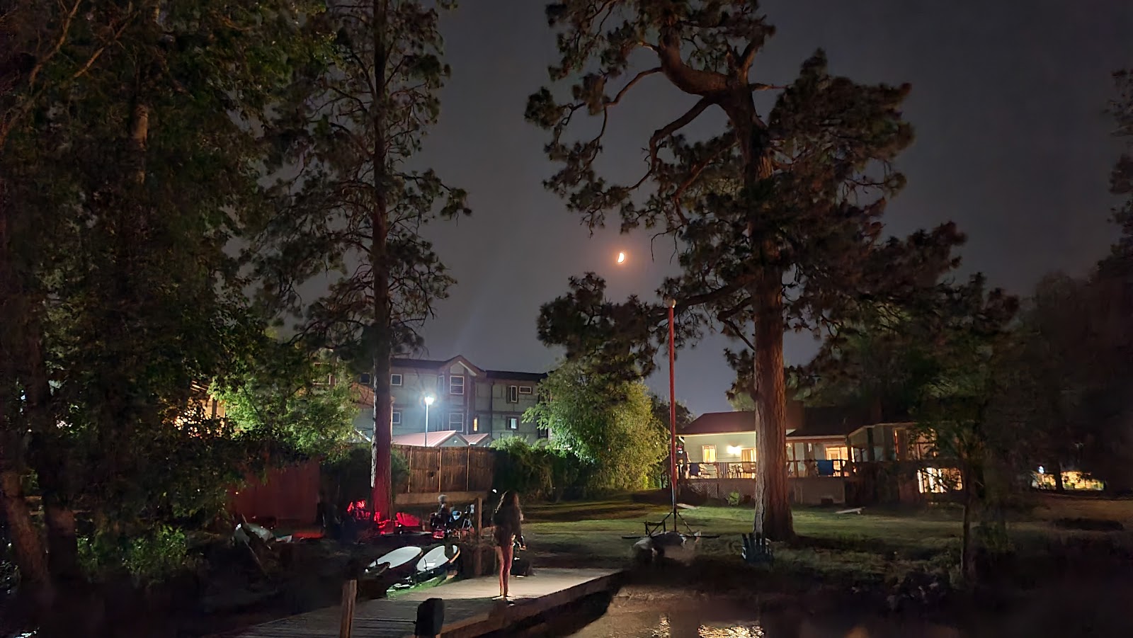Night scene at Lakeside in Glacier National Park: wooden dock, parked kayaks, tall trees, lit cabins, crescent moon.