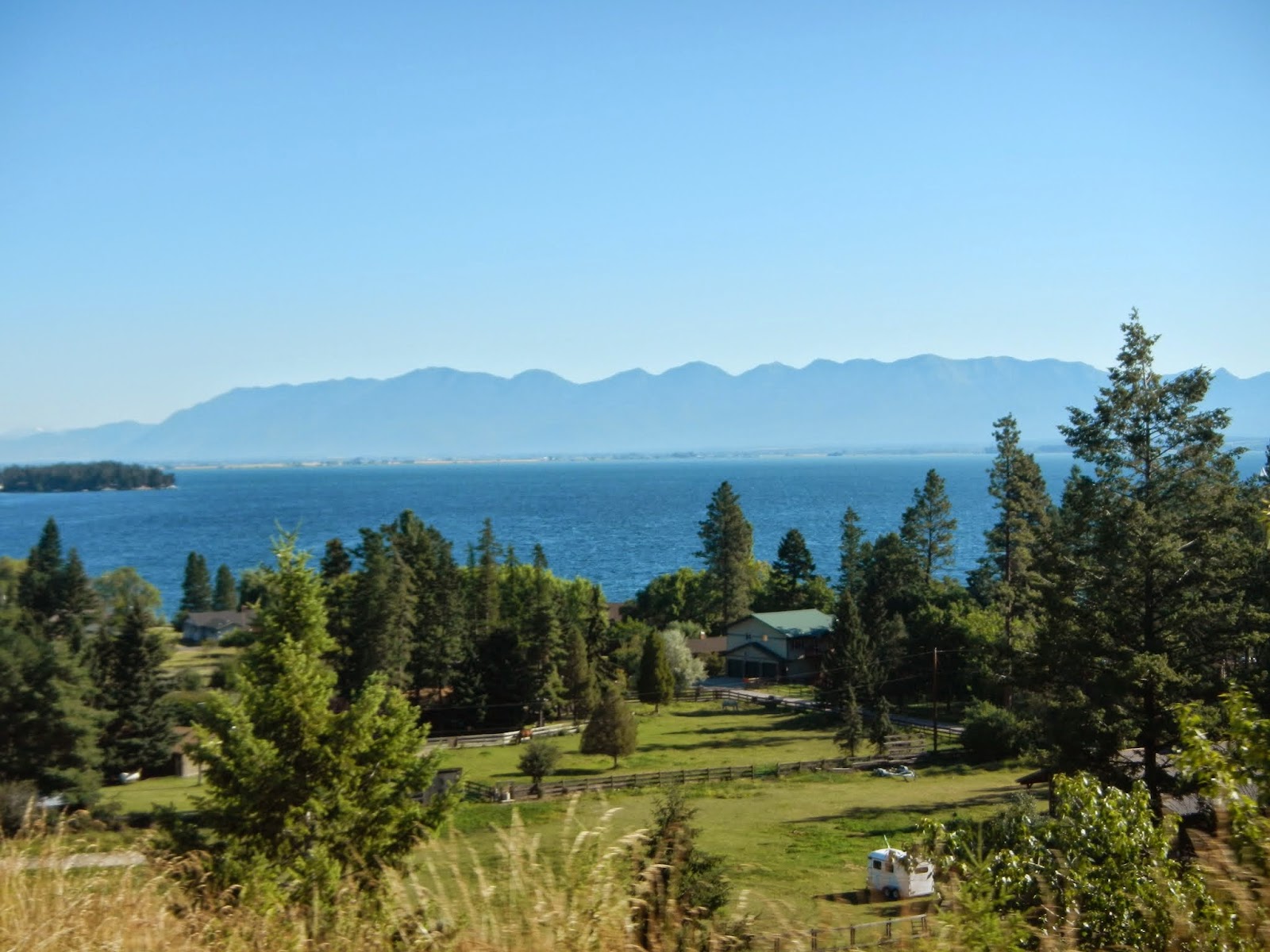 Lakeside valley in Glacier National Park with a blue lake, distant mountain range, pine trees, and scattered houses.