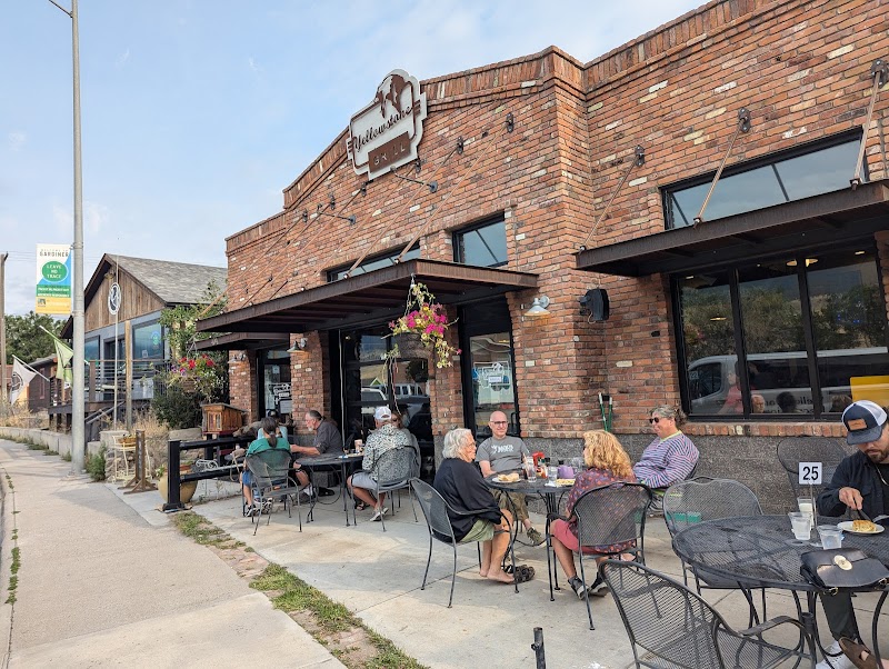 Outdoor seating at a brick restaurant with groups of people dining, hanging flower baskets, along a sidewalk in Yellowstone National Park.
