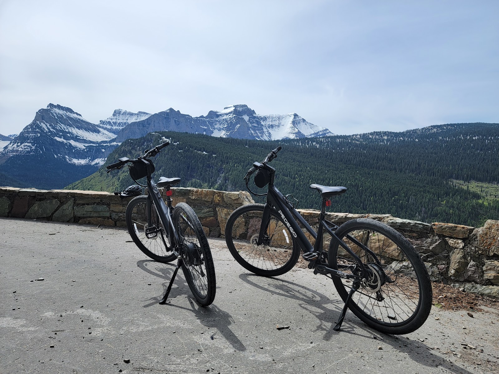 Two e-bikes rest on a paved overlook with snow-capped peaks and dense evergreen forest in Glacier National Park.