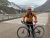 Bike rider with orange jacket and red gloves stands with a gray Specialized bike on a bridge in Glacier National Park.
