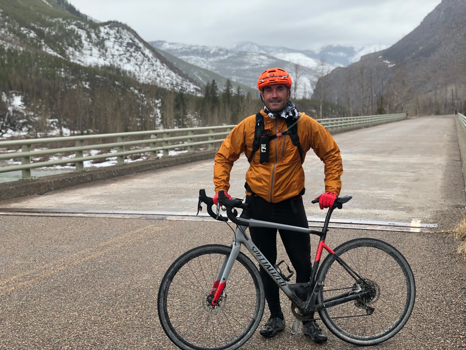 Bike rider with orange jacket and red gloves stands with a gray Specialized bike on a bridge in Glacier National Park.