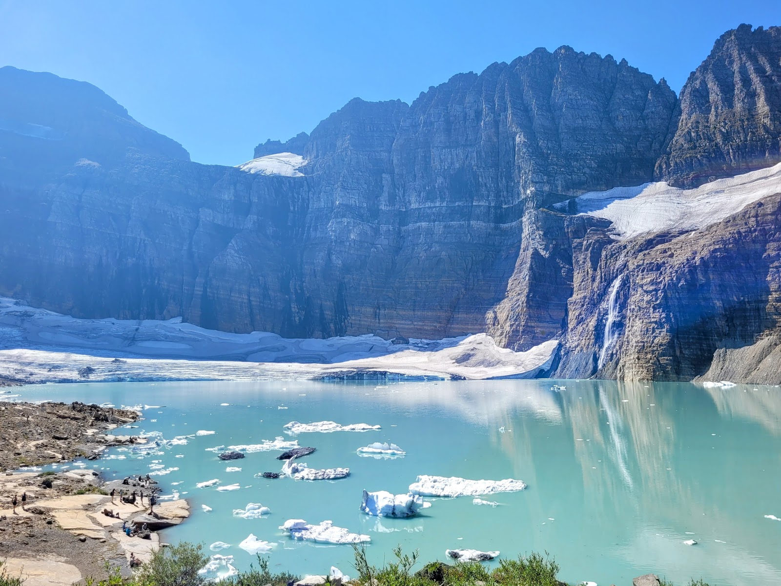 Turquoise glacier lake in Glacier National Park with floating ice, snow-dusted cliffs, and hikers along the rocky shore.