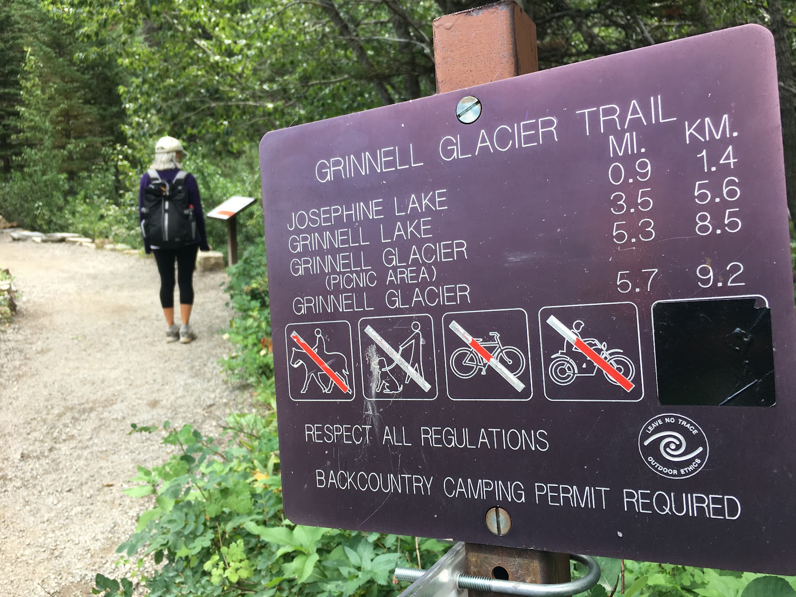 Grinnell Glacier Trailhead sign with distances and icons, a hiker on a forest trail in Glacier National Park.