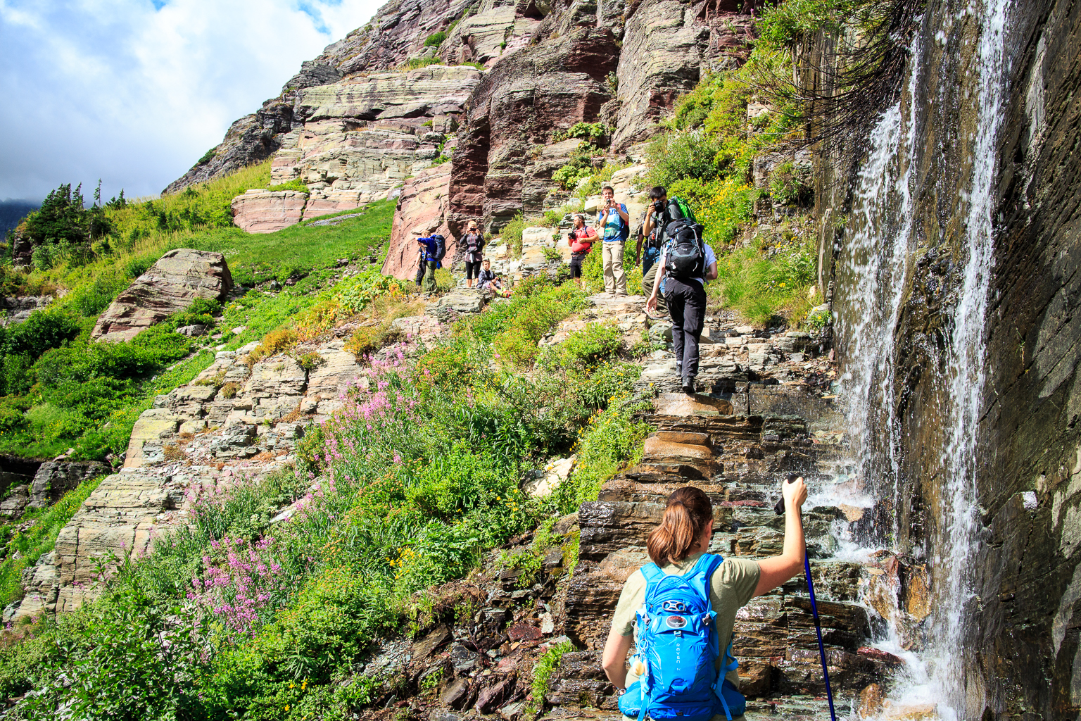 Hikers on a rocky, terraced trail at Grinnell Glacier Trailhead beside a waterfall in Glacier National Park.