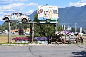 Rusty floral-painted vintage car and cartoon camper atop a wooden platform beside a flower-filled wagon in Glacier National Park.