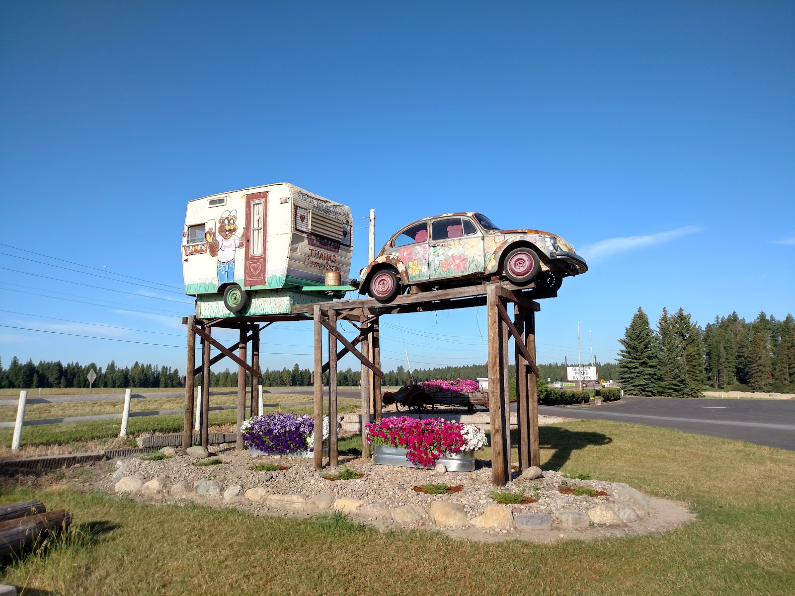 A vintage floral-painted car and cartoon trailer sit on a wooden lift above colorful flower pots, under a clear Glacier National Park sky.