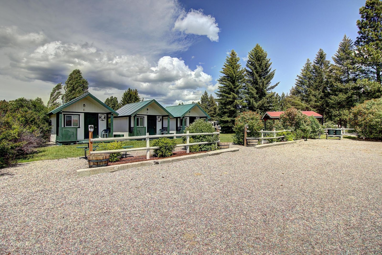 Green-roofed cabins line a gravel lot with a white fence, barrels, and shrubs in Glacier National Park, under tall evergreen trees and a partly cloudy sky.