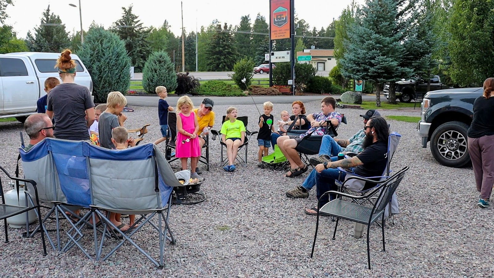 Families and kids sit in folding chairs around a campfire on a gravel lot, with trees and parked vehicles in Glacier National Park.
