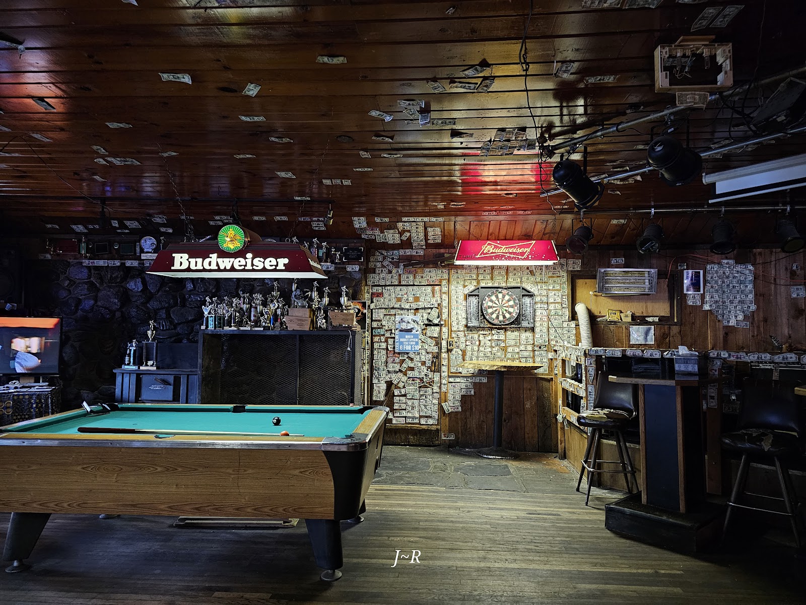 Inside a rustic Glacier National Park bar, a green felt pool table sits near money-stuck wood walls, trophies, and a central dartboard.