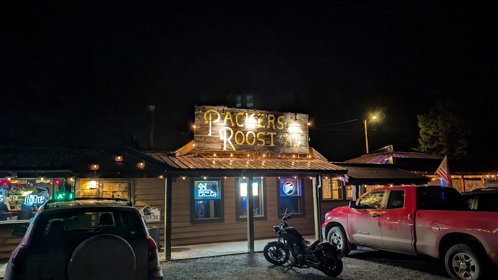 Rustic wooden restaurant lit by string lights at night in Glacier National Park, with signs, a red pickup, and a motorcycle.