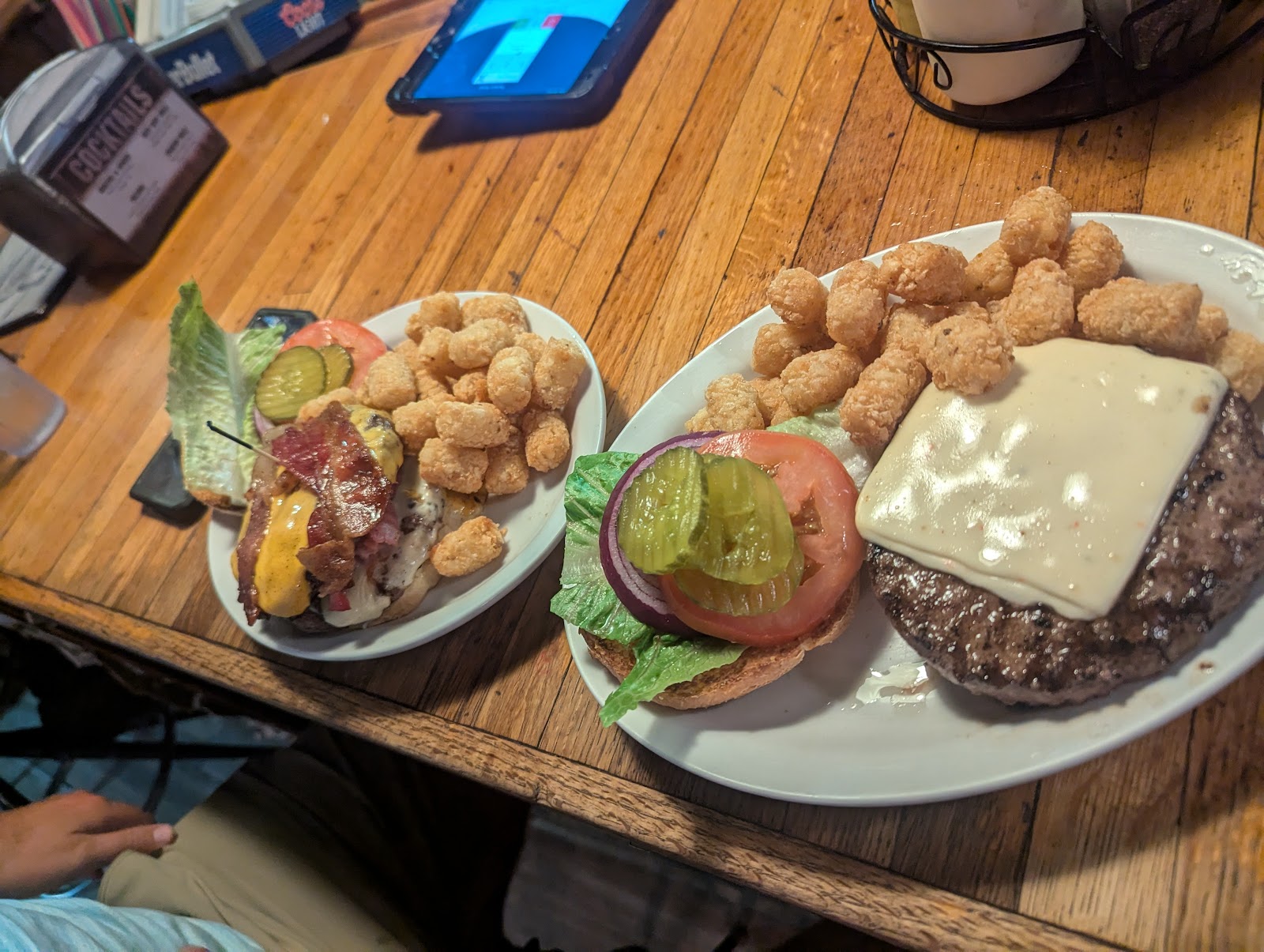 Two burger plates with cheese, bacon, lettuce, tomato and pickles, plus a side of tater tots on a wooden table in Glacier National Park.