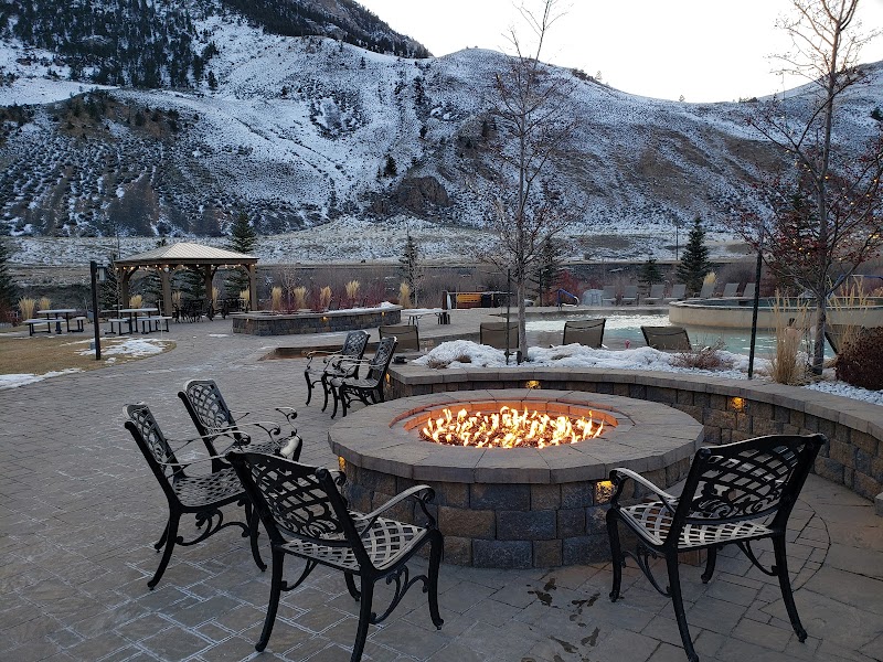 Yellowstone National Park: circular stone fire pit with bright flames, black iron chairs, and a snow-dusted mountain backdrop.