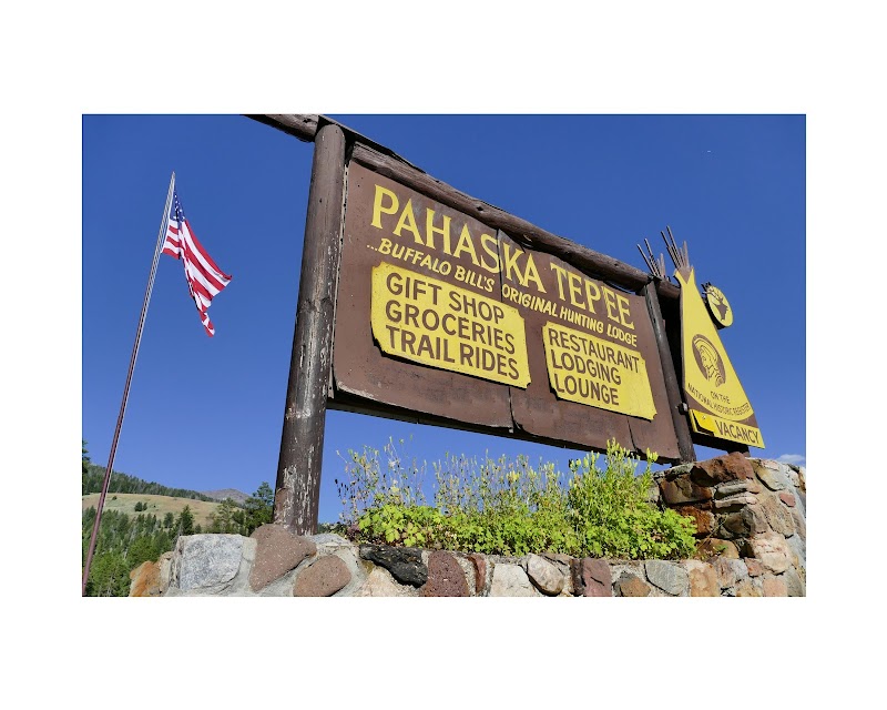Wooden sign with yellow panels advertising lodging, gifts and trail rides sits on a stone wall at Yellowstone National Park, under a clear blue sky.
