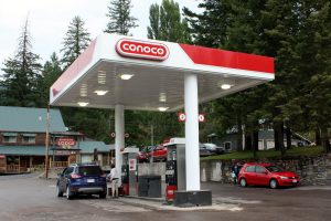 Gas station canopy and pumps at Glacier National Park, with a Conoco sign and surrounding lodge-area buildings amid tall evergreen trees.