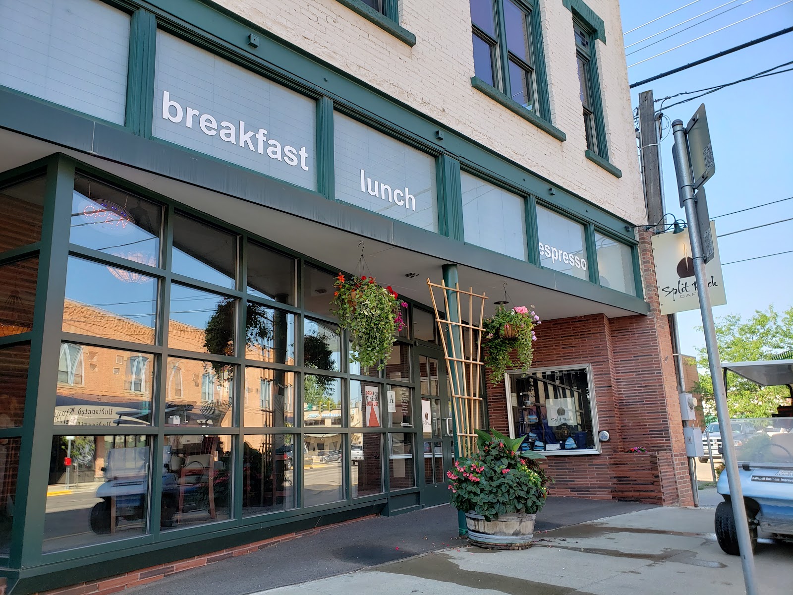 Green-framed brick storefront with large windows, hanging baskets, and potted flowers in Kalispell, Glacier National Park.