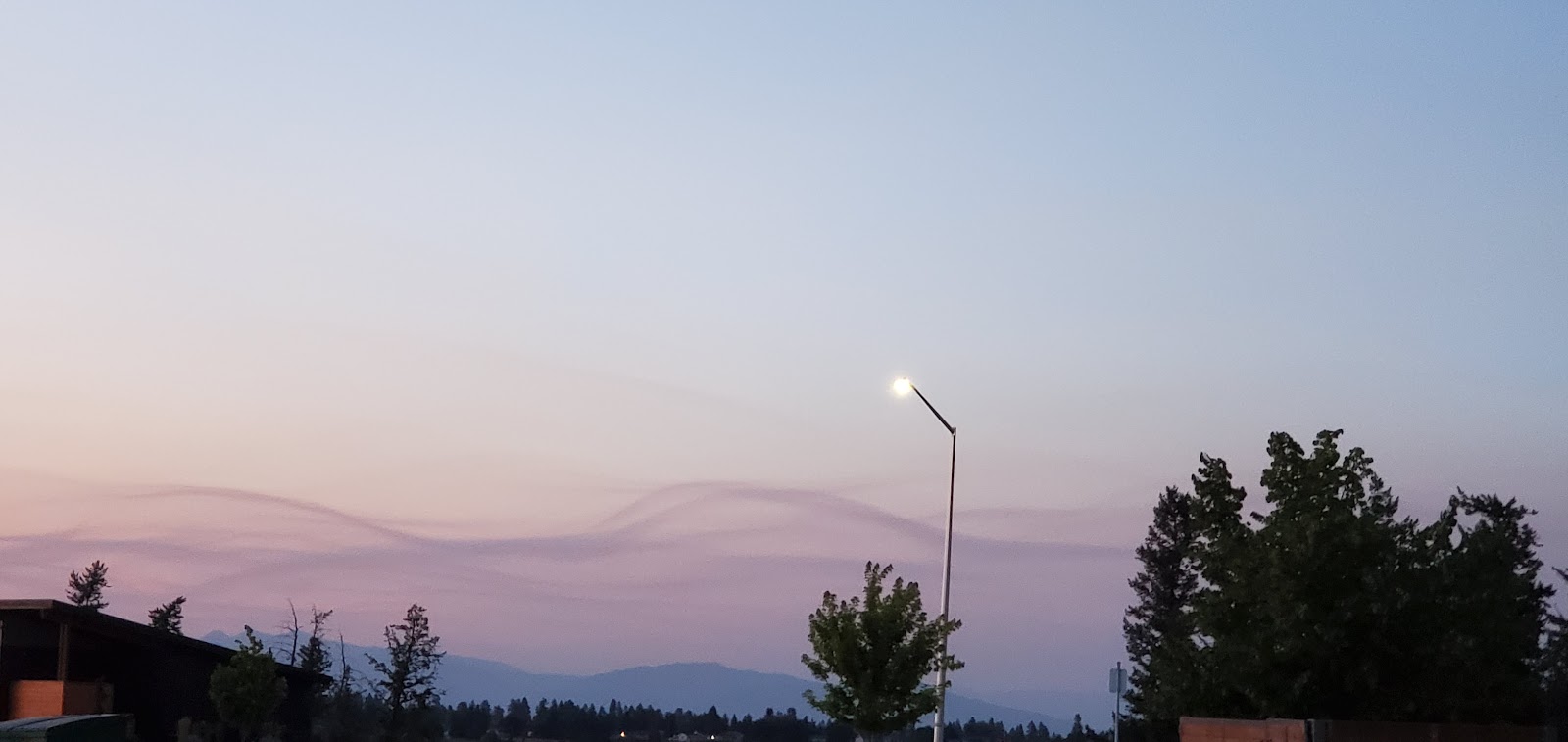 Twilight sky over a Glacier National Park area town, with a glowing streetlamp, silhouetted trees, and distant purple hills.
