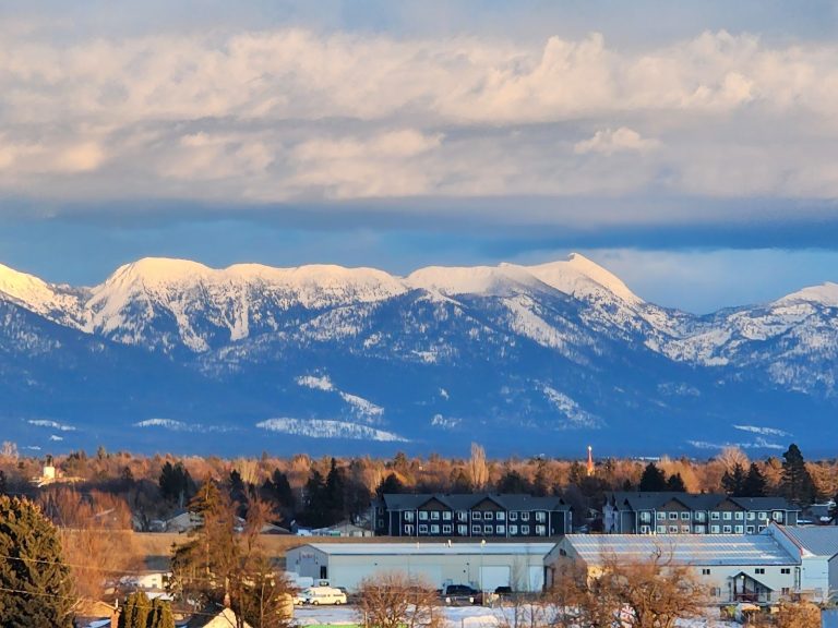 Kalispell town with snow-covered Glacier National Park peaks looming in the distance, under a pale spring sky.