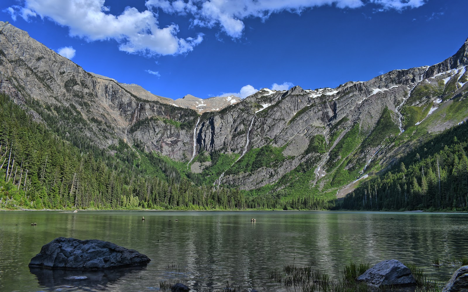 Monument Falls overlook along a tranquil lake in Glacier National Park, with jagged granite cliffs and a bright blue sky.