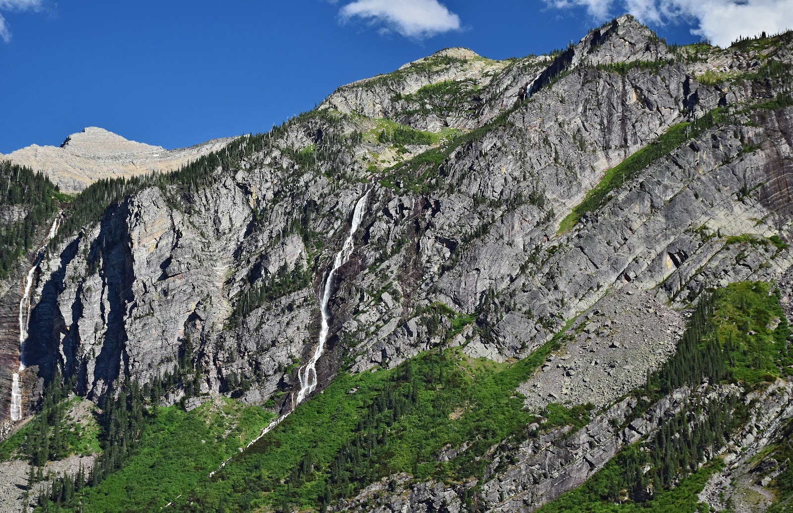 Monument Falls flows down a rugged Glacier National Park cliff, with evergreen trees and craggy rock faces.